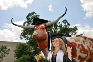 Eine Frau in einer Graduationskappe und -robe lächelt und hält einen Strauß, vor einem Denkmal, Bäumen, einem Gebäude und einem bewölkten Himmel.