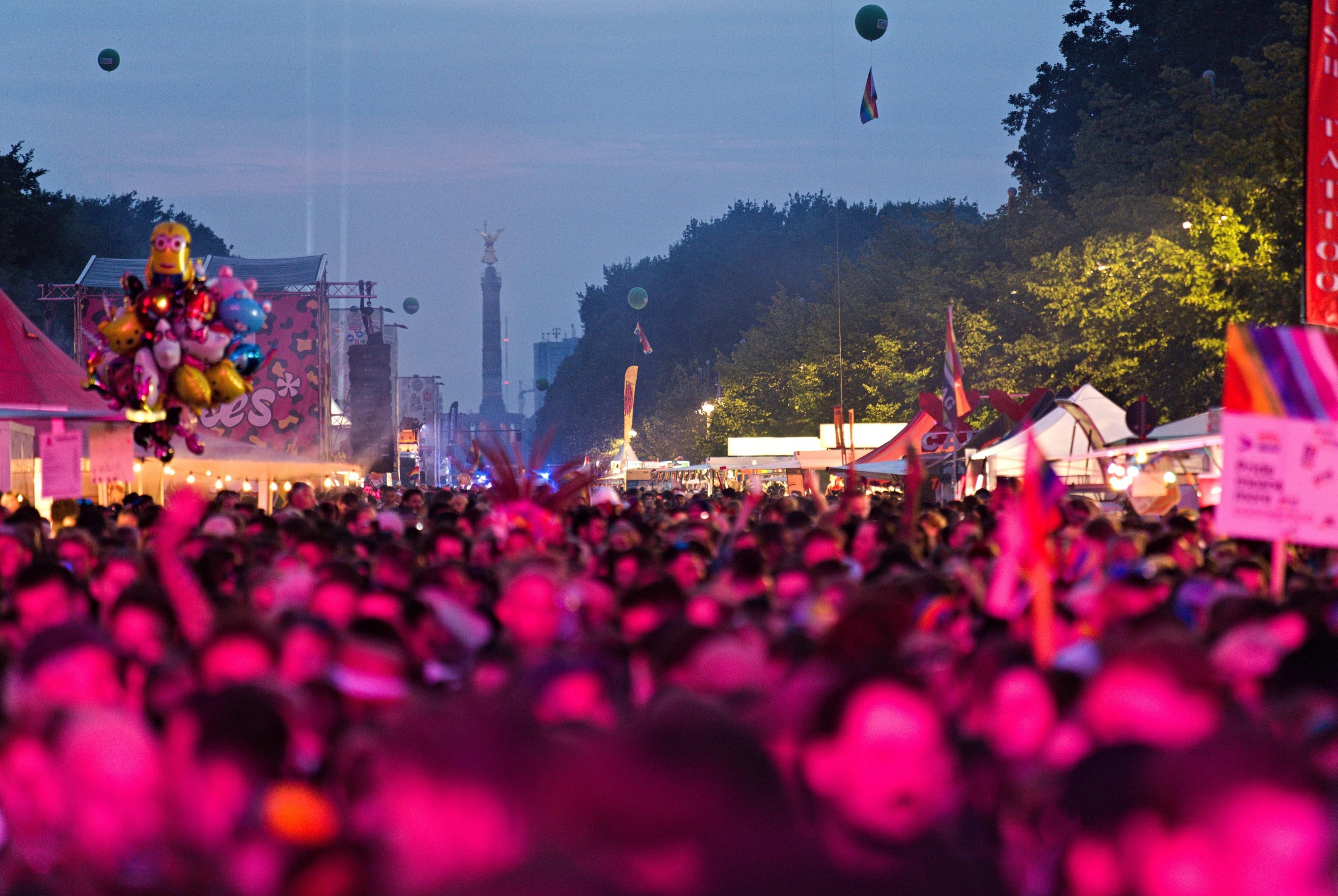Eine große Menschenmenge bei einem nächtlichen Musikfestival, beleuchtet von bunten Lichtern und Ballons, mit Zelten, Bannern, Fahnen, Bäumen, einem Turm und einem sternenklaren Himmel im Hintergrund.