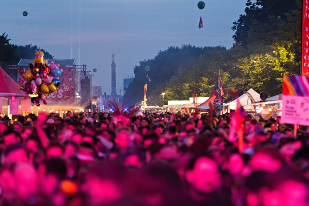 Eine große Menschenmenge bei einem nächtlichen Musikfestival, beleuchtet von bunten Lichtern und Ballons, mit Zelten, Bannern, Fahnen, Bäumen, einem Turm und einem sternenklaren Himmel im Hintergrund.