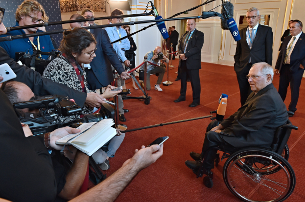 Ein Mann im Rollstuhl umgeben von Reportern mit Kameras, Mikrofonen und Handys sowie einem Banner, Scheinwerfern und einer Wand im Hintergrund.