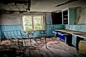 Ein zerstörter Raum mit einem Metallbett, Schränken, einem Fenster mit Bäumen und sichtbaren Wänden, Decke und Boden.