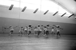 Eine Gruppe junger Mädchen in einem Tanzstudio, die sich an den Händen halten und auf dem Boden tanzen, mit einem Ball auf der rechten Seite, Lampen an der Decke und einem Objekt an der Wand im Hintergrund, dargestellt in Schwarz-Weiß.