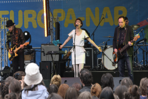 Ein Konzertauftritt mit vier Personen auf der Bühne: eine singende Frau in einem weißen Kleid in der Mitte, zwei Gitarristen links und rechts und eine Menge im Vordergrund sowie eine Wand und Lautsprecher im Hintergrund.