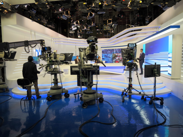 Ein TV-Studio mit mehreren Kameras auf dem Boden, einer sitzenden Person im Vordergrund und einigen stehenden Personen im Hintergrund, mit an der Decke montierten Scheinwerfern für eine Live-Broadcast-Aufnahme.