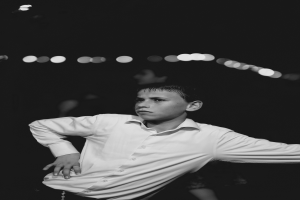 Ein junger Junge in einem weißen Hemd tanzt auf einer Tanzfläche umgeben von Menschen und Lichtern, dargestellt in Schwarz-Weiß.