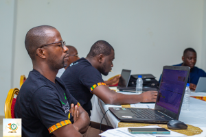 Eine Gruppe von Männern sitzt um einen Tisch mit Laptops, Wasserflaschen, Mobiltelefonen, Papieren und anderen Gegenständen. Im Hintergrund ist eine Wand mit einem Logo und Text zu sehen, der 'Nigerian Government Launches Digital Governance Programme' lautet.