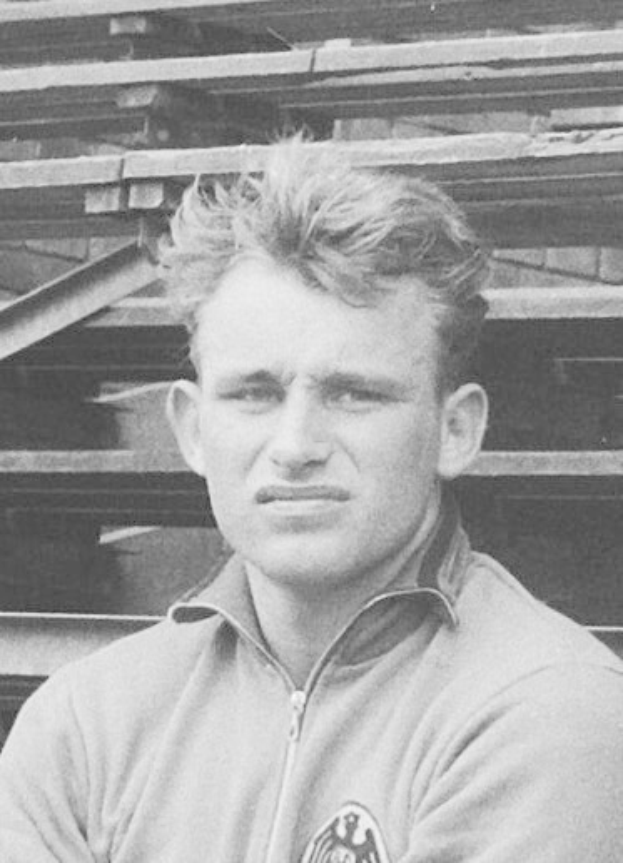 Schwarzes und weißes Foto eines Mannes in einer Jacke, der vor gestapelten Holzplanken steht, der legendäre deutsche Fußballspieler.