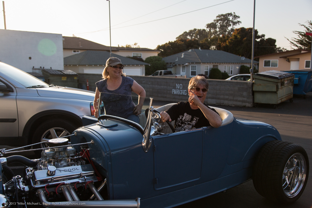 Ein sonniger Tag mit Bäumen und Häusern im Hintergrund, zwei Autos und ein Fahrzeug auf der Straße, ein Mann, der lachend auf einem Goggles trägt, und eine Frau, die Goggles und eine Kappe trägt und in ein Auto steigt.