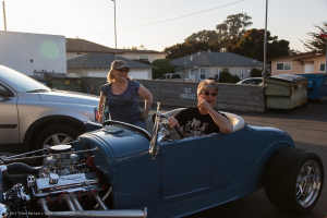 Ein sonniger Tag mit Bäumen und Häusern im Hintergrund, zwei Autos und ein Fahrzeug auf der Straße, ein Mann, der lachend auf einem Goggles trägt, und eine Frau, die Goggles und eine Kappe trägt und in ein Auto steigt.