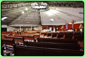 Ein Innenblick in eine Kirche mit Holzliegen und sitzenden Menschen, einer Statue und Blumensträußen im Hintergrund und einer Decke darüber.