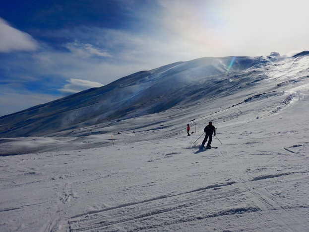 Eine Person, die eine schneebedeckte Piste mit Bergen im Hintergrund hinunterskifährt, trägt einen Helm, Handschuhe und Skischuhe und hält Ski-Stöcke in der Hand, unter einem bewölkten Himmel mit glitzerndem Schnee in der Sonne.