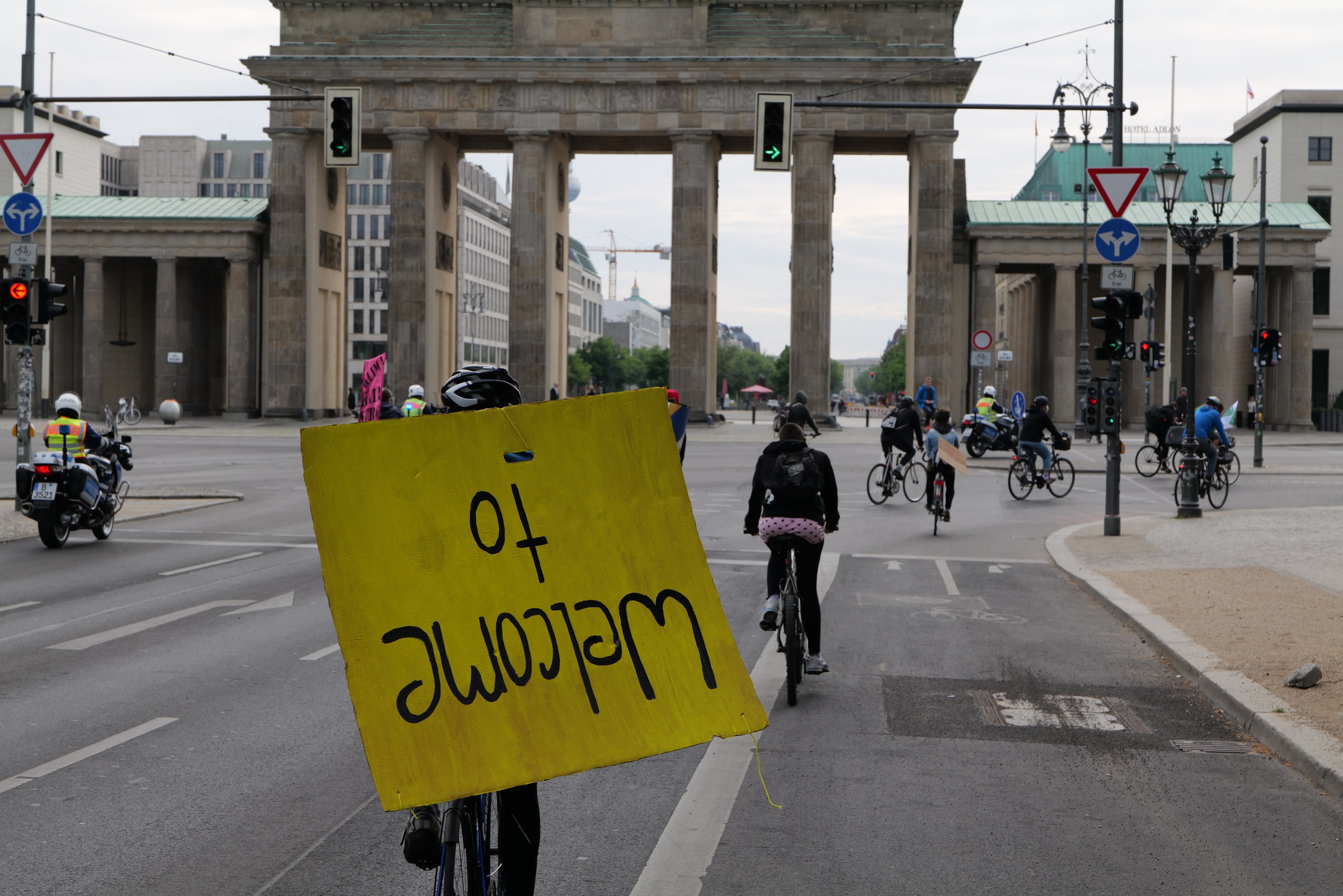 Gruppe von Radfahrern am Brandenburger Tor in Berlin vorbei, einer hält ein gelbes Schild, mit Laternen, Ampeln, Gebäuden, Bäumen und einem klaren Himmel im Hintergrund.