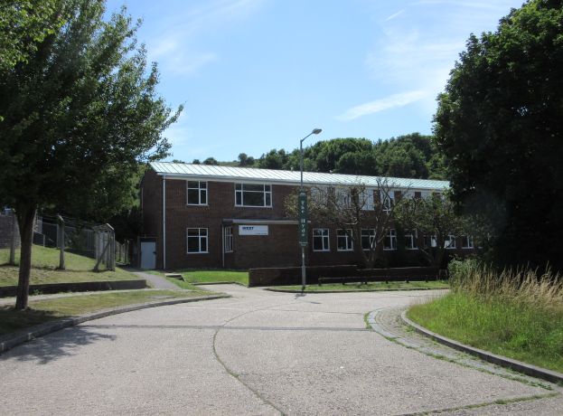 Ein Schulgebäude mit Fenstern, einem Straßenmast, einem Schild, einer Baumgruppe, Gras, einem Zaun und einer bewölkten Himmel, das inmitten eines Parks umgeben von saftigem Grün liegt.