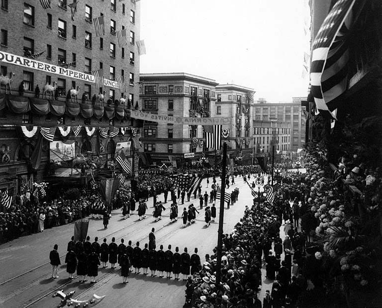 Eine große Menschenmenge marschiert auf einer Straße in New York City während eines Fourth of July-Umzugs, viele tragen Kappen und halten Fahnen, während andere an den Seiten stehen; Fahnen an Stangen und Bäumen säumen die Straße, und Gebäude mit Fenstern und Text auf einer Fassade sind im Hintergrund zu sehen.