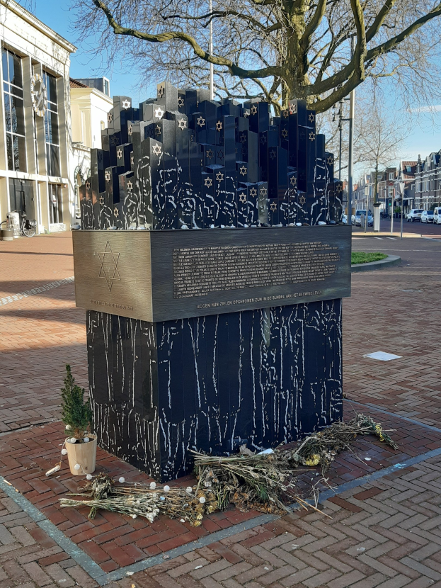 Denkmal in einem Stadtplatz für Holocaust-Opfer, mit einem Baum davor, einem Topfgewächs, verstreuten trockenen Blättern und städtischem Hintergrund mit Gebäuden, Fahrzeugen, Pfählen, Bäumen und Himmel.