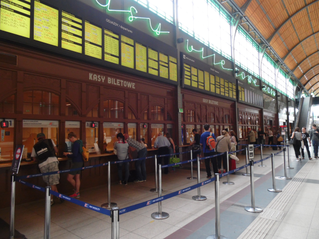 Menschen in einer Warteschlange an einem Bahnhofsschalter mit Pfählen, Bändern, Informationsschildern, Deckenleuchten und Treppen mit Geländern im Hintergrund.