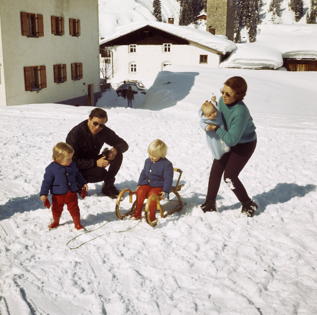 Eine Familie von vier Personen, die im Schnee spielt, wobei der Mann auf einem Schlitten sitzt und die Frau ein Baby hält, vor einer Kulisse von Häusern, Bäumen und einem klaren blauen Himmel.