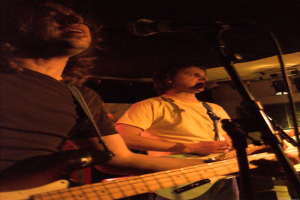 Eine Person in einem weißen Hemd singt in ein Mikrofon und spielt eine grüne Gitarre, während sie neben einer anderen Person in einem schwarzen Hemd und Brille steht, die ebenfalls singt und eine schwarze Gitarre spielt, mit Scheinwerfern und anderen Instrumenten im Hintergrund.