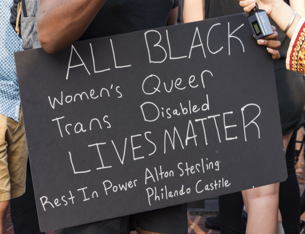 Eine Gruppe von Menschen, die zusammenstehen, wobei eine Person in der Mitte ein Schild hält, auf dem steht: "All Black Women's Queer Trans-Disabled Lives Matter", und eine andere Person ein Objekt hält.