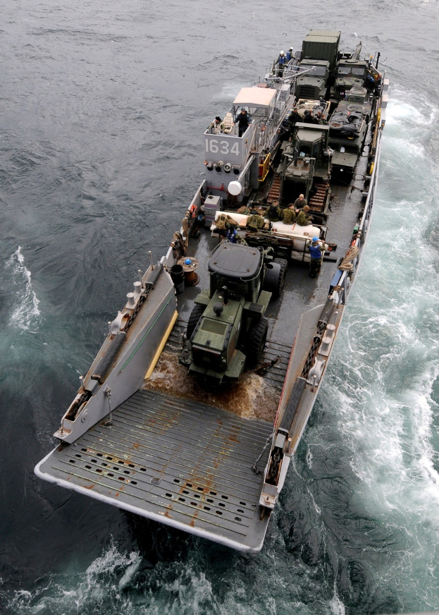 Luftaufnahme eines großen Bootes auf dem Meer mit einem Militärfahrzeug darauf, umgeben von Menschen und Ausrüstung, mit einem Wasserzeichen unten.