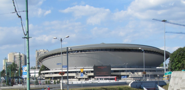 Großes Stadion mit Fahrzeugen, Fußgängern und städtischer Infrastruktur vorne, vor einem bewölkten Himmel.