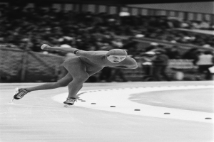 Ein Schwarz-Weiß-Foto eines Eisschnellläufers in Aktion auf dem Eis, der eine Mütze und Eislaufschuhe trägt, mit ein paar unscharfen Menschen und einem Zaun im Hintergrund.