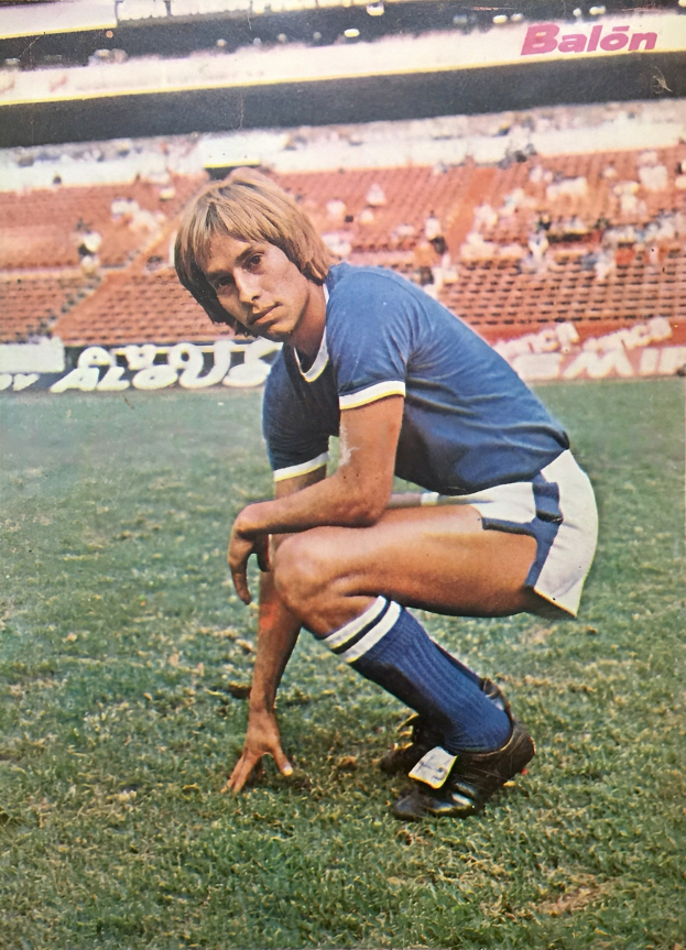 Ein Mann kniet auf einem Fußballfeld umgeben von Menschen auf Stühlen und Brettern mit Text, trägt Schuhe.