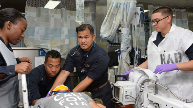 Eine Gruppe von vier Personen um einen Mann auf einer Trage in einem Krankenhauszimmer mit medizinischer Ausrüstung, Vorhängen, einer Kochsalzflasche und Deckenlampen, wahrscheinlich in einem Notfallraum der US-Küstenwache.