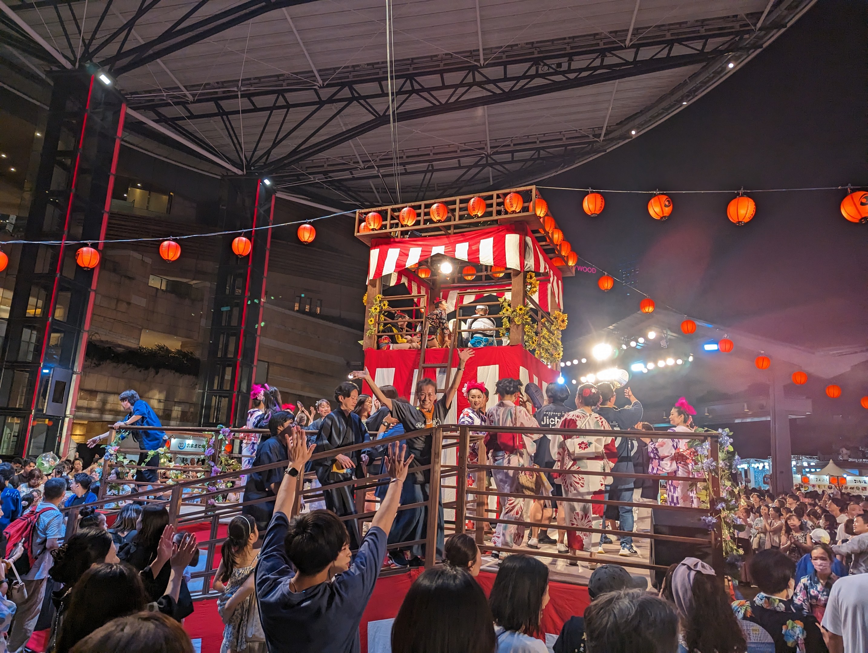 Eine Menschenmenge auf einer beleuchteten Bühne mit Laternen, einige halten Kameras, umgeben von einem Geländer, mit Bäumen und einem blauen Himmel im Hintergrund bei Nacht, wahrscheinlich während eines japanischen Festivals.