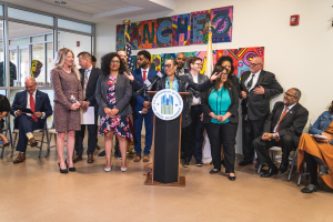 Gruppe von Menschen vor einem Rednerpult mit Mikrofon und einer Tafel mit der Aufschrift "Sancho-Stadtrat tritt dem Stadtrat bei", mit sitzenden und stehenden Teilnehmern, Flaggen, Feuerlöscher und Deckenleuchten im Hintergrund.