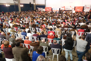 Ein volles Stadion mit vielen Sitzplätzen und Stehplätzen, eine Bühne mit Personen in der Nähe eines Podiums, Banner und Lichter.