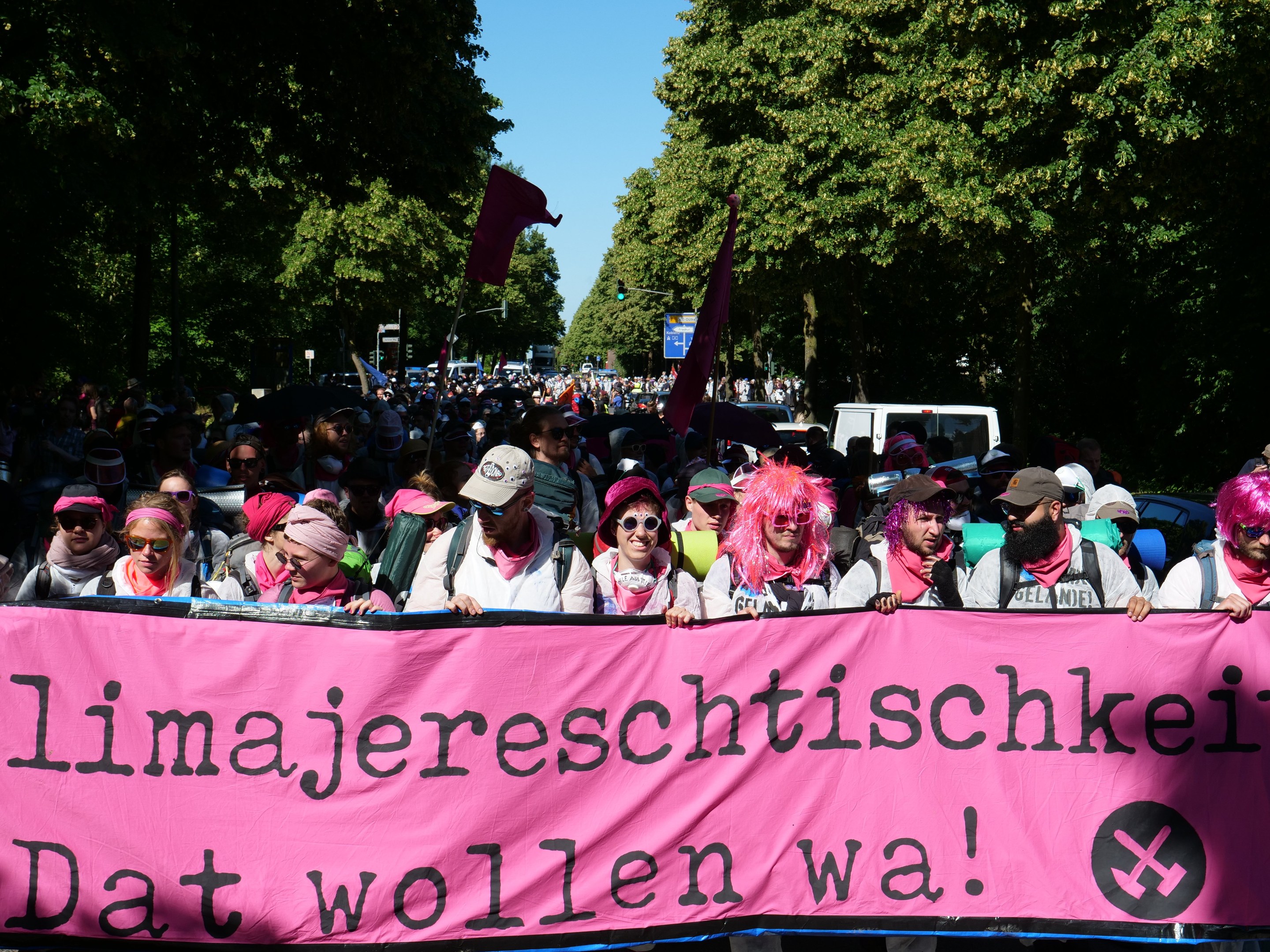 Eine Gruppe von Menschen marschiert auf einer Straße, hält ein pinkes Banner mit der Aufschrift "Women's March in Berlin" und trägt teilweise Mützen und Goggles sowie Fahnen; Bäume, Fahrzeuge und Himmel sind im Hintergrund zu sehen.