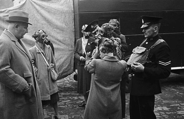 Schwarzes und weißes Bild von Menschen in Mänteln und Mützen mit Gasmasken, die in einer Reihe vor einem Zelt mit einem Fahrzeug im Hintergrund stehen.