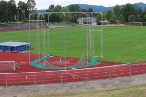 Fußballfeld mit einem Netz darüber, von einem Metallzaun umgeben, mit einem zentralen Trampolin, Gras, Bäumen, Häusern, Pfosten, einer Treppe und einem bewölkten Himmel.