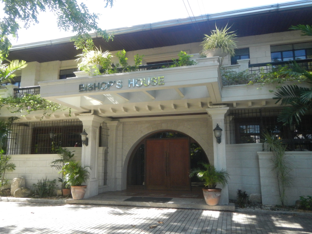 Das Bischofshaus in Manila, Philippinen, ein Gebäude mit Fenstern, einer Tür, einer Namensschilder, Topfpflanzen, nahen Bäumen, verstreuten Steinen und einem bewölkten Himmel.