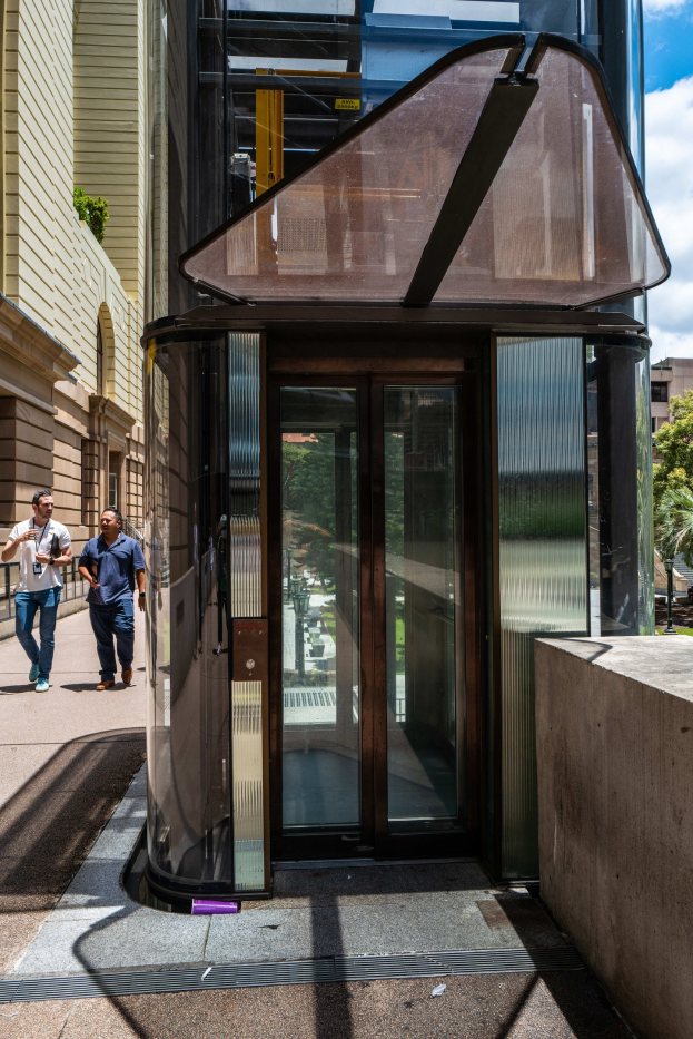 Glaselevator in einer Stadtstraße umgeben von Passanten, mit einer Wand auf der rechten Seite und Gebäuden, Bäumen und einem bewölkten Himmel im Hintergrund.