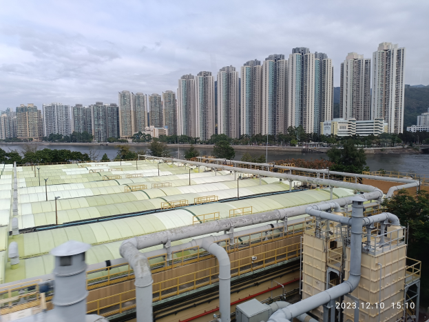 Ein Stadtpanoramablick von einem Gebäude aus, mit Gebäuden, Bäumen, Pfählen, Wasser im Vordergrund und einem bewölkten Himmel im Hintergrund, mit einem Wasserzeichen, das eine Kläranlage in Hong Kong anzeigt.