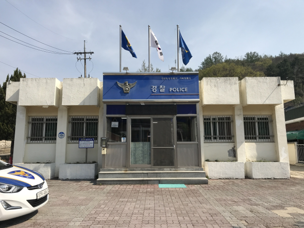 Eine Polizeiwache mit einem geparkten Auto, Fenstern, einer Tür und einer Tafel mit Text auf dem Gebäude, Flaggen im Hintergrund, einem Strommast mit Drähten, bewölktem Himmel und entfernten Bäumen.
