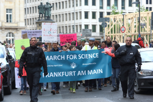 Eine Gruppe von Menschen marschiert mit einer "March for Science Frankfurt am Main"-Plakette die Straße entlang, mit Autos, Gebäuden, Statuen, Laternen, Schildern und Bäumen im Hintergrund.