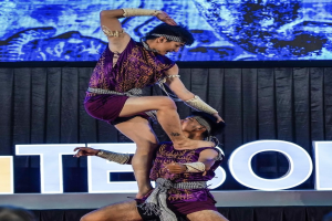 Zwei Performer in purpurfarbenen Kleidern führen eine akrobatische Hebung auf der Bühne vor einem Publikum aus, mit einem schwarzen Vorhang und einer Leinwand im Hintergrund.