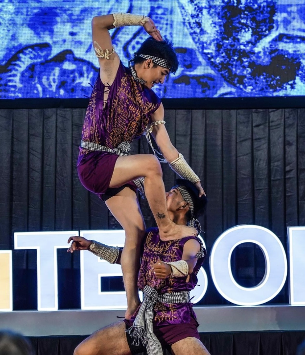 Zwei Performer in purpurfarbenen Kleidern führen eine akrobatische Hebung auf der Bühne vor einem Publikum aus, mit einem schwarzen Vorhang und einer Leinwand im Hintergrund.