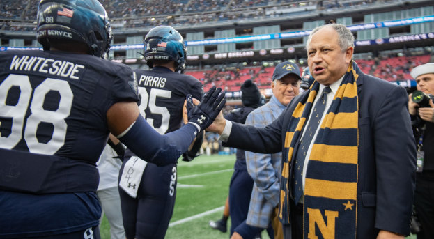 Ein Mann im Anzug schüttelt einem Fussballspieler auf einem Feld die Hand, umgeben von einer Menge, während eine Person mit einer Kamera und einer Mütze rechts steht; im Hintergrund ist ein Stadion mit Zuschauern und Schildern mit der Aufschrift "Notre Dame Football Coach Mike McCarthy and the Fighting Irish" zu sehen.