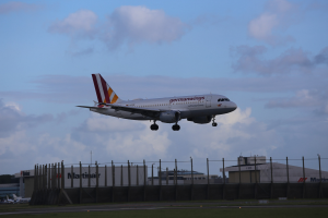 Germanwings Airbus A320-200 beim Abheben vom Frankfurter Flughafen mit Gras darunter, einem Zaun im Vordergrund, Gebäuden und Bäumen im Hintergrund und einer bewölkten Himmel darüber.