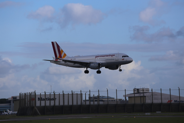 Germanwings Airbus A320-200 beim Abheben vom Frankfurter Flughafen mit Gras darunter, einem Zaun im Vordergrund, Gebäuden und Bäumen im Hintergrund und einer bewölkten Himmel darüber.