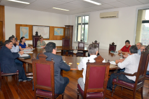 Eine Gruppe von Menschen sitzt an einem Tisch mit Papieren und Gläsern, ein Klimagerät, ein Anschlagbrett und Karten an der Wand im Hintergrund.