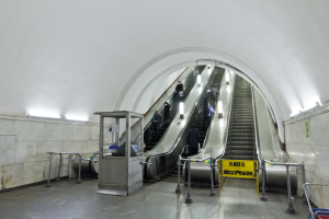 Eine Rolltreppe in einer U-Bahn-Station mit Menschen, Geländern, Texttafeln, Wandlampen und einem Einkaufswagen unten.