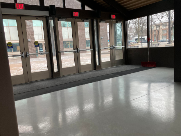 Großer Raum mit hoher Decke, mehreren Fenstern und Türen, einem Besen auf dem Boden, Schildern, einem sichtbaren Dach, umliegenden Bäumen, geparkten Fahrzeugen, nahegelegenen Gebäuden und einem bewölkten Himmel, was auf einen gewerblichen Bodenreinigungsdienst hinweist.