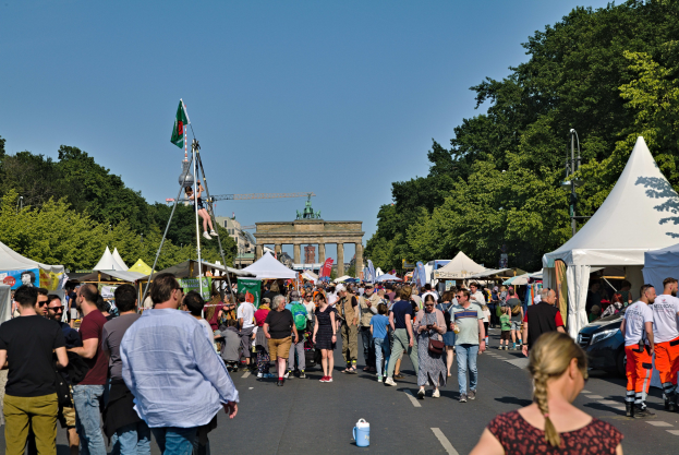 Eine Menschenmenge, die eine Straße mit Zelten, Fahrzeugen und Bäumen entlanggeht, mit einem Tor und einem klaren blauen Himmel im Hintergrund und Fahnenmasten auf der linken Seite.
