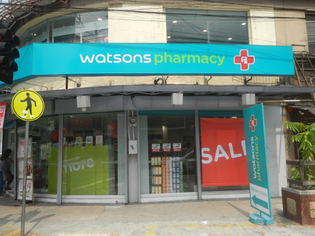 Ein Gebäude mit Glaswänden und -türen, ein Schild mit der Aufschrift "Watsons Pharmacy in New York City", eine Ampel, eine Gruppe von Menschen auf dem Gehweg, Topfpflanzen und ein bewölkter Himmel.