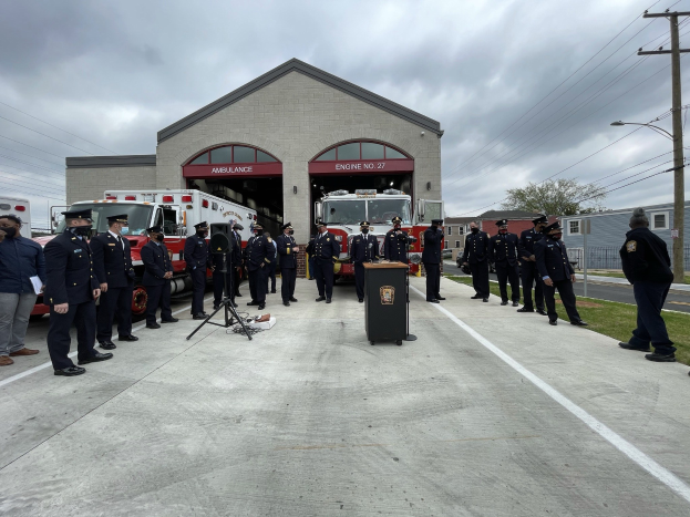 Gruppe von Menschen in Mützen vor einem Löschfahrzeug auf einem Podium während einer Feuerwache-Einweihungszeremonie, mit Gebäuden, Bäumen, Strommasten, Laternen und Gras im Hintergrund unter einem bewölkten Himmel.